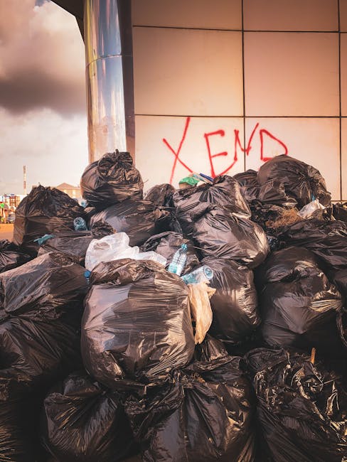 A large pile of black rubbish bags filled with waste is stacked against a modern, reflective building with a tiled exterior wall. The bags are tightly packed and some are partially torn, revealing pla
